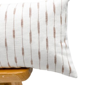 Pu&ograve; includere: Un cuscino rettangolare bianco con un motivo a righe verticali marroni. Il cuscino &egrave; appoggiato su uno sgabello di legno. Il cuscino ha un aspetto strutturato e una palette di colori morbidi e neutri.