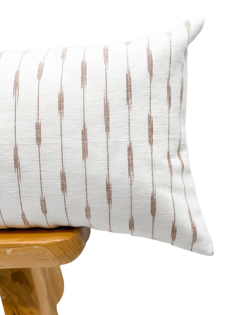 Pu&ograve; includere: Un cuscino rettangolare bianco con un motivo a righe verticali marroni. Il cuscino &egrave; appoggiato su uno sgabello di legno. Il cuscino ha un aspetto strutturato e una palette di colori morbidi e neutri.