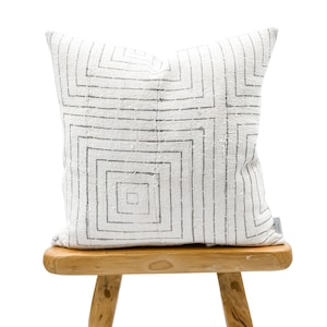 May include: Square decorative pillow with a geometric gray pattern on a white background. The pillow is placed on a small, light-colored wooden stool. The pillow's design features a series of nested squares.