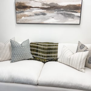May include: A white couch with six throw pillows, a green and black plaid throw blanket, and a framed abstract painting on the wall behind the couch.