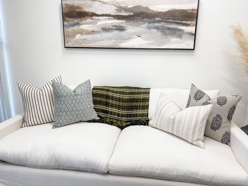 May include: A white couch with six throw pillows, a green and black plaid throw blanket, and a framed abstract painting on the wall behind the couch.