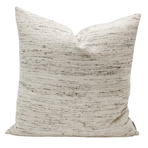 May include: A square decorative pillow with a textured, off-white fabric. The pillow has a woven appearance with dark brown and gray flecks throughout. The pillow is a neutral color and would complement various home decor styles.
