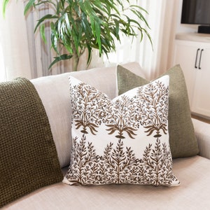 May include: A decorative pillow with a white background and a brown floral pattern. The pillow is on a light-colored sofa, with a green throw blanket and a green pillow on either side. The room has a neutral color palette.