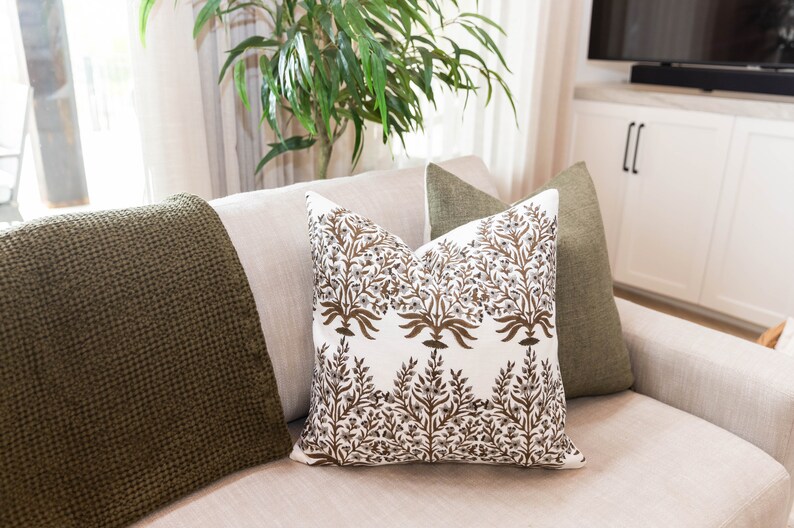 May include: A decorative pillow with a white background and a brown floral pattern. The pillow is on a light-colored sofa, with a green throw blanket and a green pillow on either side. The room has a neutral color palette.