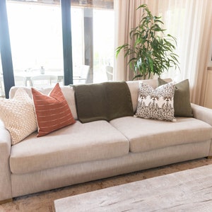 May include: A beige sofa with several decorative pillows and a green throw blanket. The pillows are in various colors and patterns, including cream, rust, and a floral design. The sofa is in a well-lit living room setting.