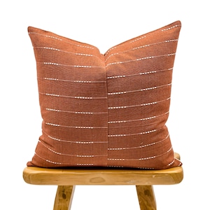 Op de afbeelding: Een vierkant kussen in roestbruin met horizontale witte stippellijnen. Het kussen is gemaakt van textiel met textuur en ligt op een kleine houten kruk. Het ontwerp van het kussen is eenvoudig en elegant.