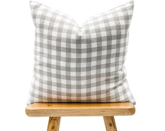 May include: A square pillow with a gray and white gingham pattern. The pillow is resting on a small, light-colored wooden stool. The pillow has a soft, textured appearance.