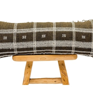 May include: A long, rectangular brown and white patterned pillow with fringe on the ends. The pillow is sitting on a wooden stool.