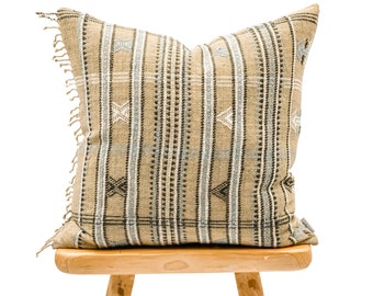 May include: A beige and blue patterned cushion with fringe trim sits on a wooden stool.