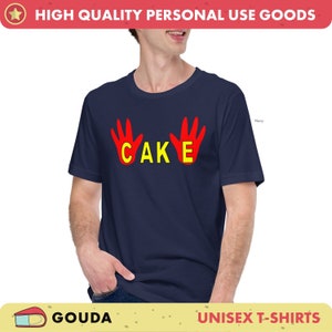 May include: A navy blue unisex t-shirt featuring the word "CAKE" in yellow, flanked by red handprints. The short-sleeved shirt is suitable for everyday wear. The text "HIGH QUALITY PERSONAL USE GOODS" is at the top.