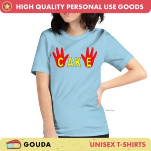 May include: Pale blue unisex t-shirt featuring the word "CAKE" in yellow, with red handprints on either side. The shirt is labelled "Ocean Blue" and is part of a "UNISEX T-SHIRTS" collection. The brand name "GOUDA" is also visible.