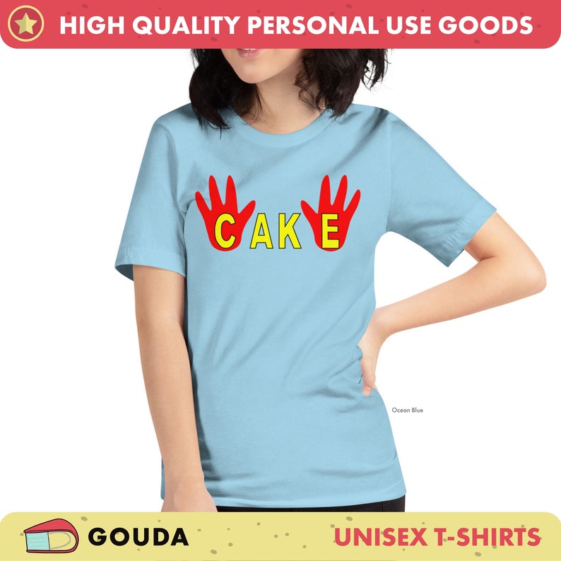May include: Pale blue unisex t-shirt featuring the word "CAKE" in yellow, with red handprints on either side. The shirt is labelled "Ocean Blue" and is part of a "UNISEX T-SHIRTS" collection. The brand name "GOUDA" is also visible.