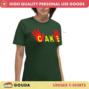 May include: A forest green unisex t-shirt featuring the word "CAKE" in yellow, with red hand graphics. The t-shirt has a crew neck and short sleeves.
