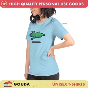 May include: A light blue unisex t-shirt with a pixelated spaceship graphic in green, blue, and red colors. The text "Ocean Blue" is visible in the top right corner of the image.
