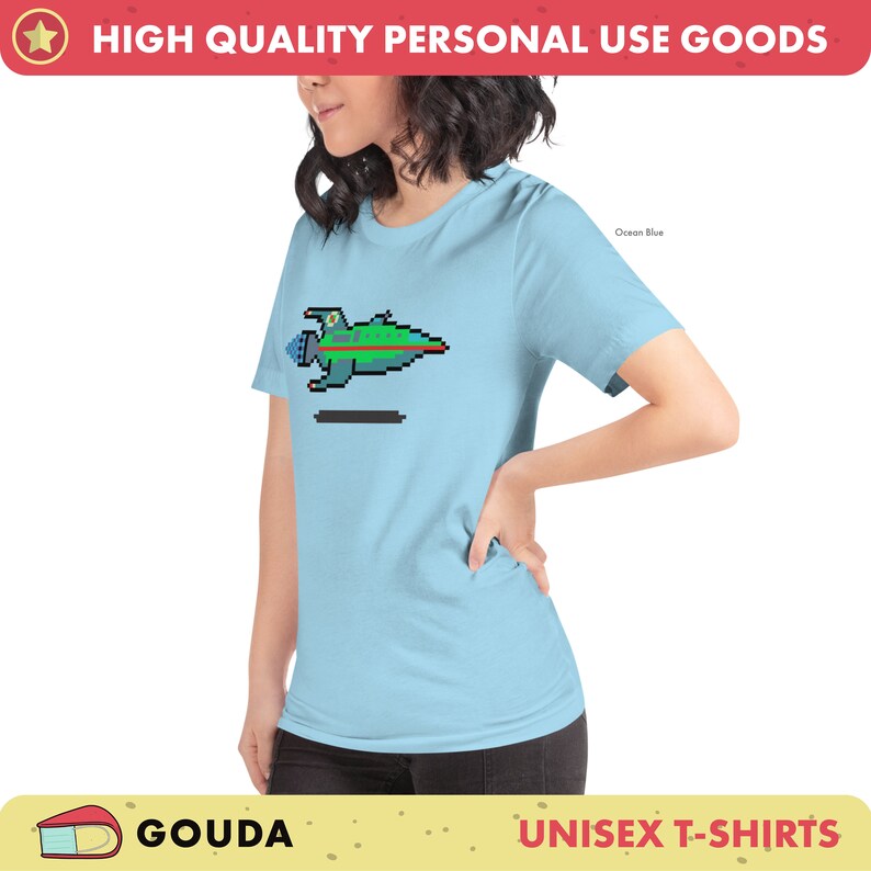 May include: A light blue unisex t-shirt with a pixelated spaceship graphic in green, blue, and red colors. The text "Ocean Blue" is visible in the top right corner of the image.