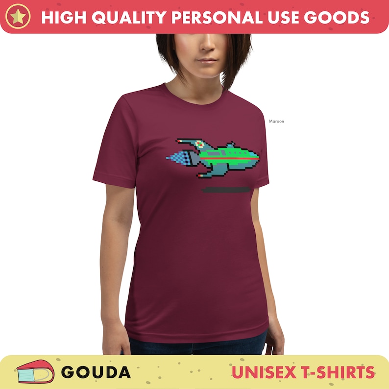 May include: A maroon unisex t-shirt featuring a pixelated spaceship design. The spaceship is green, blue, and white with a yellow flame coming out of the back. The text "Maroon" is visible in the top right corner of the image.