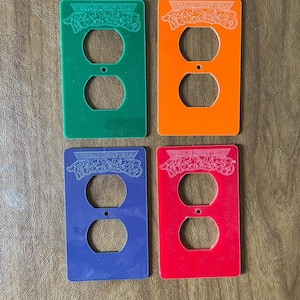 May include: Four colorful Teenage Mutant Ninja Turtles themed outlet covers. The covers are green, orange, blue, and red. Each cover has two circular openings and a small centered hole. The text "Teenage Mutant Ninja Turtles" is etched on each cover.