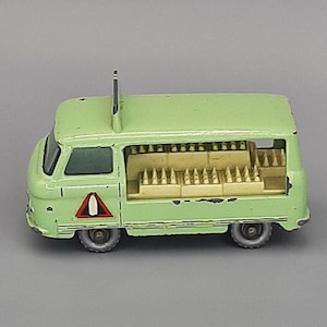 Puede incluir: Una furgoneta de juguete vintage de color verde claro con un interior detallado y una pequeña antena. La furgoneta presenta una señal de peligro en el lateral y se encuentra sobre un fondo gris. El juguete muestra signos de desgaste.