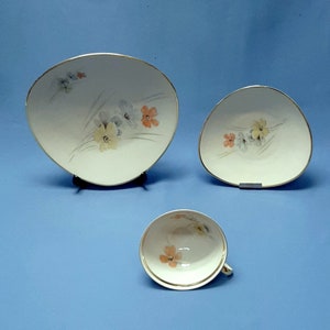 May include: A three-piece porcelain tea set with a floral design. The set includes a teacup, and two irregularly shaped plates. The pieces are white with a gold trim and feature a delicate pattern of yellow, orange, and blue flowers.