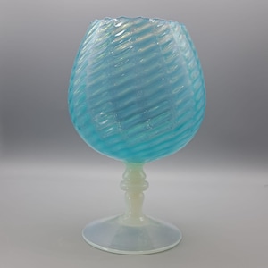 Op de afbeelding: Een grote, opalescerende glazen kelk met een brede, afgeronde kom en een korte steel. De kom heeft een wervelend, getextureerd patroon in lichtblauw en wit. De voet en steel zijn crèmewit van kleur.