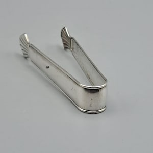 Può includere: Una pinza per zucchero color argento con un design curvo. Le pinze hanno una base arrotondata e estremità decorative a forma di ventaglio. L'immagine è scattata su uno sfondo grigio chiaro.