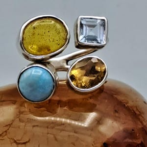 Könnte beinhalten: Ein Silberring mit vier Edelsteinen: ein gelbes Oval, ein quadratischer hellblauer Stein, ein gelb-oranges Oval und ein runder hellblauer Stein. Der Ring hat ein modernes Design, wobei die Steine in einzelnen Fassungen gefasst sind.