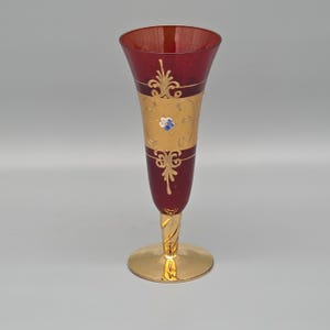 Op de afbeelding: Een sierlijke glazen vaas in dieprood met gouden details. De vaas heeft een uitlopende bovenkant, een gouden band en decoratieve bloemmotieven. De voet en steel zijn goudkleurig, met een gedraaide steel. Hoogte ongeveer 25 cm.