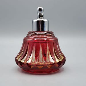 May include: A red glass perfume bottle with a silver-colored metal top. The bottle has a decorative, fluted design with gold accents. The top is a shiny, cylindrical shape with a small, rounded knob.