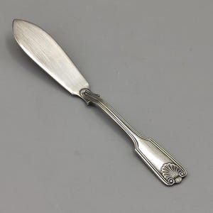 Pode incluir: Uma faca de manteiga prateada com uma lâmina arredondada e um cabo decorativo. A pega apresenta um desenho de concha na extremidade e uma série de linhas verticais. A faca é feita de metal brilhante.