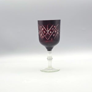 Può includere: Un calice rosso rubino con stelo e base trasparenti. Il bicchiere presenta un motivo intagliato sulla coppa. Lo stelo è trasparente e presenta un design decorativo. Il bicchiere è su sfondo bianco.