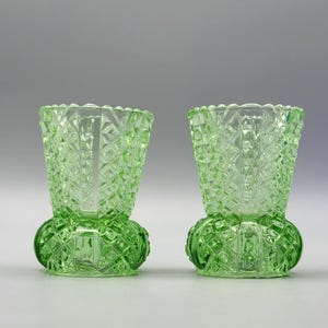 Puede incluir: Dos jarrones de vidrio verde con un patrón de diamantes y una base redondeada. Los jarrones tienen una parte superior acampanada y una superficie texturizada, mostrando una estética vintage. Los jarrones se exhiben sobre un fondo gris claro.