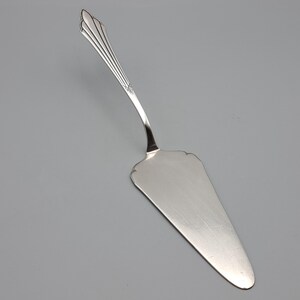 May include: A silver-colored cake server with a decorative handle. The handle has a fan-like design at the top and a curved shape. The serving blade is triangular with a scalloped edge.