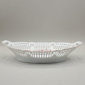 Könnte beinhalten: Weißes Porzellan-Serviertablett mit durchbrochenem Design. Die ovale Schale hat ein zartes Blumenmuster in der Mitte und einen goldenen Rand. Die Schale hat zwei Griffe und eignet sich zum Servieren von Obst oder Gebäck.