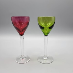 Könnte beinhalten: Zwei Weingläser mit Stiel. Ein Glas hat eine tiefrote Schale, das andere eine grüne Schale. Beide Gläser haben klare Stiele und kreisförmige Basen. Die Gläser stehen vor einem schlichten, hellgrauen Hintergrund.