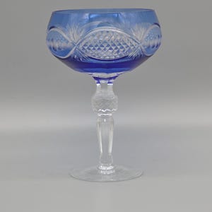 Könnte beinhalten: Ein klares Kristall-Weinglas mit einer blauen Schale und einem klaren Stiel. Die Schale hat ein rauten- und fächerförmiges Design. Der Stiel hat einen dekorativen, bauchigen Abschnitt. Das Glas steht auf einem klaren, kreisförmigen Fuß.