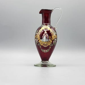 Puede incluir: Una jarra de vidrio rojo rubí con un asa de vidrio transparente. La jarra presenta un diseño ovalado pintado con una mujer con un vestido, rodeada de filigranas doradas y detalles florales. La base es de vidrio transparente.