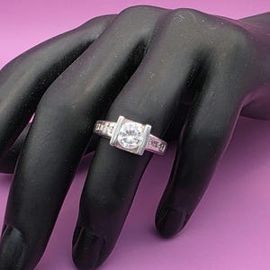 Può includere: Un anello d'argento con una grande gemma rotonda e trasparente incastonata in una lunetta quadrata. La fascia presenta pietre più piccole e trasparenti. L'anello è mostrato su una mano nera su sfondo viola.