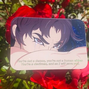 Op de afbeelding: Een sticker met een tekening van een persoon met donker haar en ogen, die huilt. De achtergrond is donkerblauw met witte sterren. De tekst op de sticker luidt: "You're not a demon, you're not a human either. You're a devilman, and so I will save you."