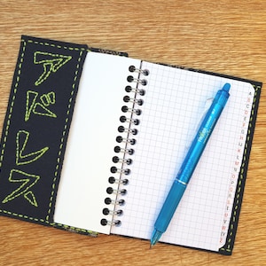 Può includere: Un quaderno nero con un disegno ricamato verde sulla copertina. Il quaderno ha una rilegatura a spirale ed è aperto su una pagina bianca con una penna blu sopra. Il quaderno ha un indice alfabetico sul lato destro.