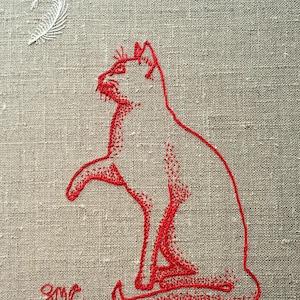 Puede incluir: Obra de arte bordada con un gato rojo en posición sentada, con una pata levantada. Una pluma blanca está encima del gato sobre un fondo de lino neutro. Las iniciales "SMC" están bordadas debajo.