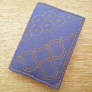 Può includere: Una copertina per libro in tessuto viola con un motivo geometrico cucito con filo dorato.