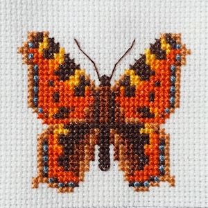 May include: A cross-stitch embroidery of a butterfly with orange, brown, and blue wings.
