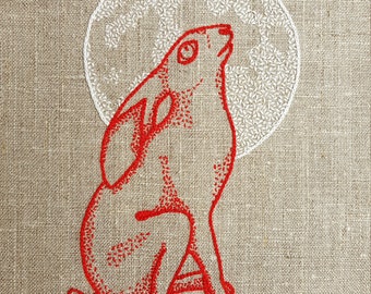 May include: Red embroidered design of a rabbit sitting up and looking at a white embroidered moon on a beige linen background. The design is signed "smce".