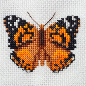 Peut inclure: Une broderie au point de croix représentant un papillon aux ailes orange, brun, noir et blanc.
