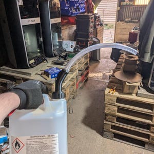 Könnte beinhalten: Ein durchsichtiger Plastikbehälter mit einem schwarzen Ausguss und einem weißen Etikett mit der Aufschrift "ISOPROPANOL 99,9%". Der Behälter wird von einer behandschuhten Hand gehalten. Der Behälter befindet sich vor einer Holzpalette.