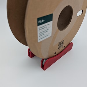 Op de afbeelding: Een bruine 3D-printer filamentspoel met een rode spoelhouder. De spoel heeft een label met "PLA+". De tekst "3D PRINTER FILAMENT" is op de spoel gedrukt.