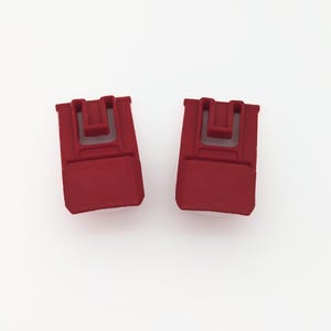 May include: Two red plastic clips with a U-shaped opening. The clips are designed to hold items together.