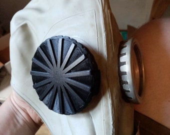 May include: A vintage gas mask with a cream-colored rubber facepiece and a black, circular filter. The mask has a metal canister and is held by a person. The mask is designed to protect the wearer from hazardous substances.