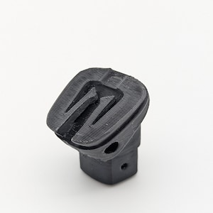 May include: A black, 3D-printed object with a unique, geometric design. The object has a rectangular base and a top section with a series of parallel grooves. The overall shape is angular and modern, with a textured surface.