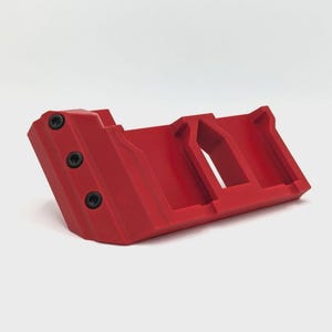 May include: A bright red, angular object with a geometric design. The object has two black screws on one side and a series of rectangular cutouts. The item is made of a solid material and is set against a white background.